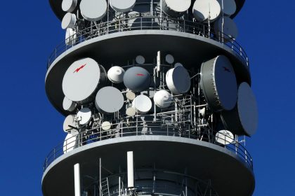 Detalhe de uma torre de telecomunicações com várias antenas e pratos de satélite contra o céu.