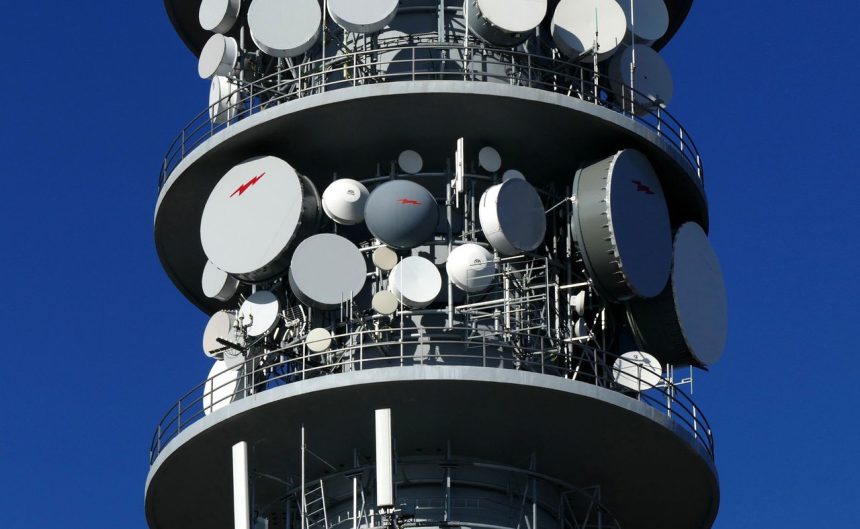 Detalhe de uma torre de telecomunicações com várias antenas e pratos de satélite contra o céu.